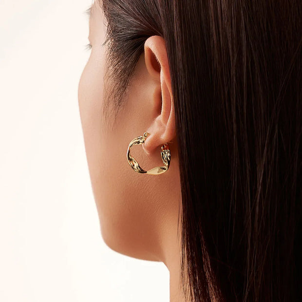 A profile view of a model wearing the sculptural 14K gold-plated irregular wavy hoop earrings.