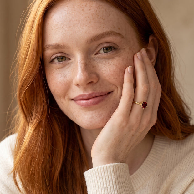 Model with copper hair wearing the 14K gold-plated 925 silver solitaire ring with a garnet-red CZ in six prongs, hand at the cheek.